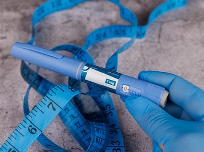 Eine Hand mit blauen Handschuhen pr&auml;sentiert einen Ozempic- Pen nebst einem Ma&szlig;band