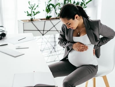Eine schwangere Frau sitzt im Office, h&auml;lt ihren Bauch mit den H&auml;nden und ertr&auml;gt Schmerzen